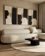 A modern living room featuring a three-panel abstract wall art set with black, beige, and gold brushstrokes in thin gold frames, hung above a textured beige boucle sofa.