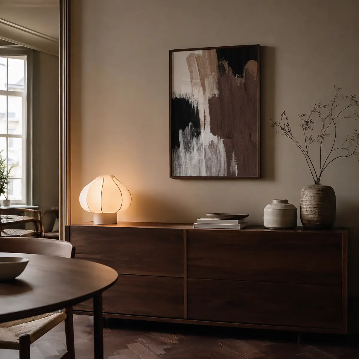 A framed abstract wall art print with black, brown, and white brushstrokes hangs above a dark wood sideboard in a warm, modern interior. 