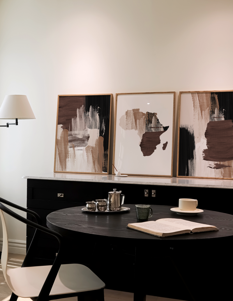 A set of three abstract strokes chocolate wall art prints displayed on a wall above a round table with a lamp, books, and a cup on it, in a room with dark flooring and white walls.
