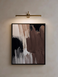 Abstract art piece with brown, black, and white brushstrokes in a black frame, illuminated by a wall-mounted light fixture.