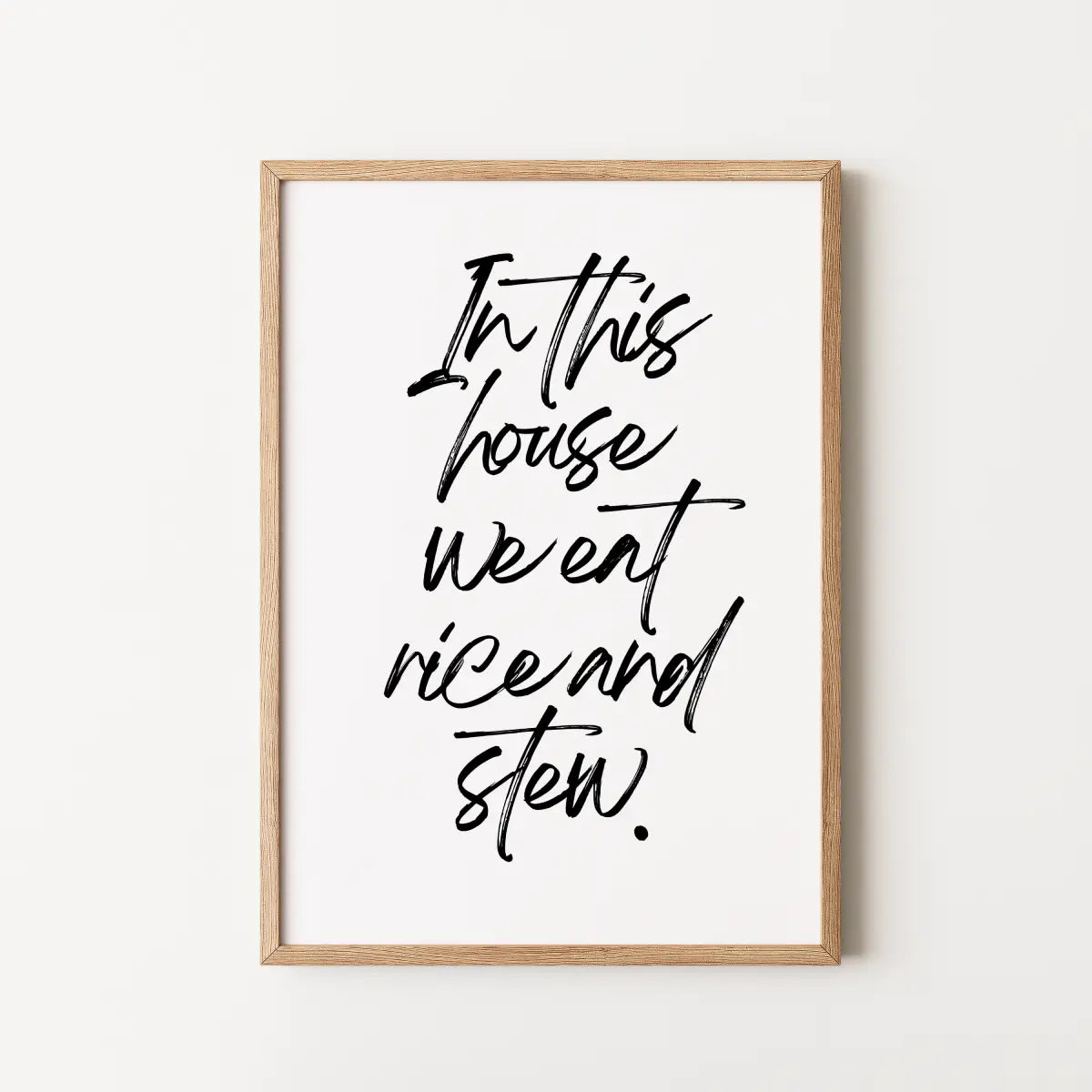 Framed typographic wall art with black handwritten text reading 'In this house we eat rice and stew.' 