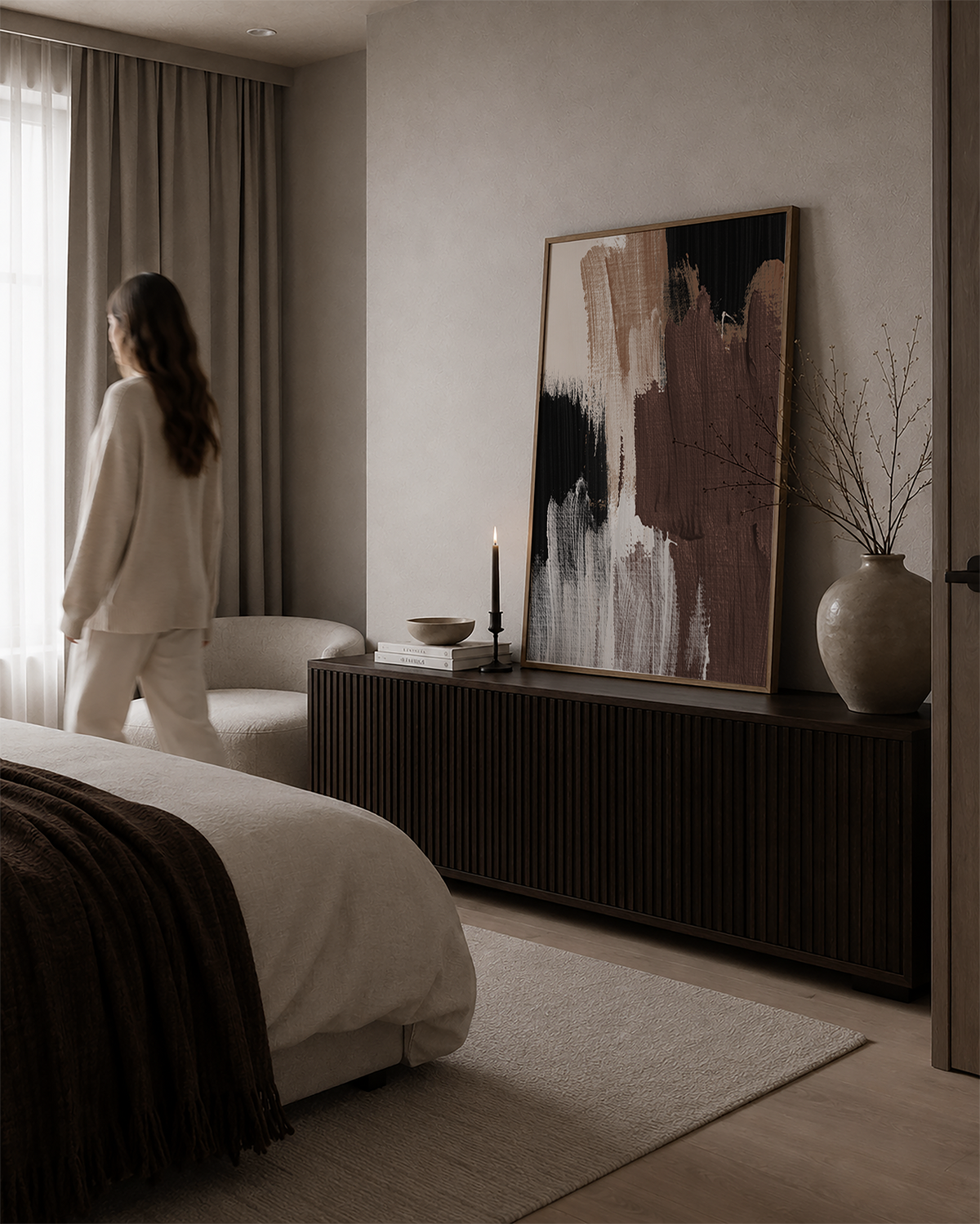 A woman in a cream pajama set stands by a window in a modern, minimalist bedroom with brown artwork.