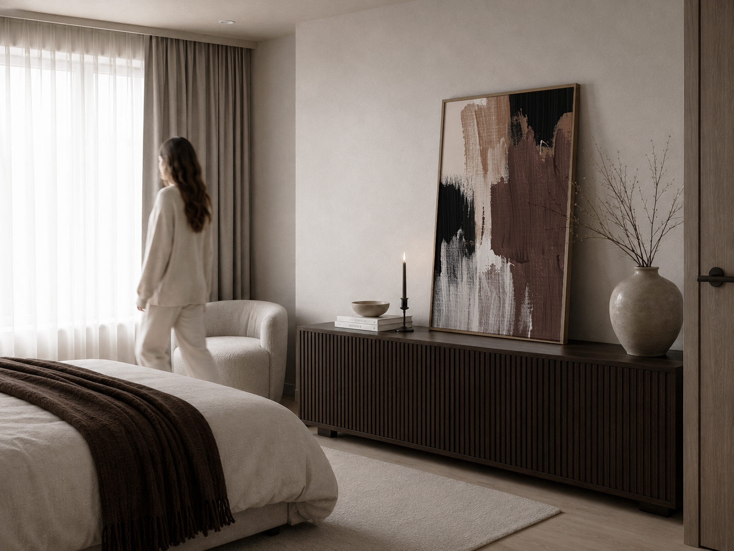 A woman in a cream pajama set stands by a window in a modern, neutral-toned bedroom with brown wall art.