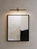 Abstract neutral beige art print in a black frame on a wall with a gold light fixture.