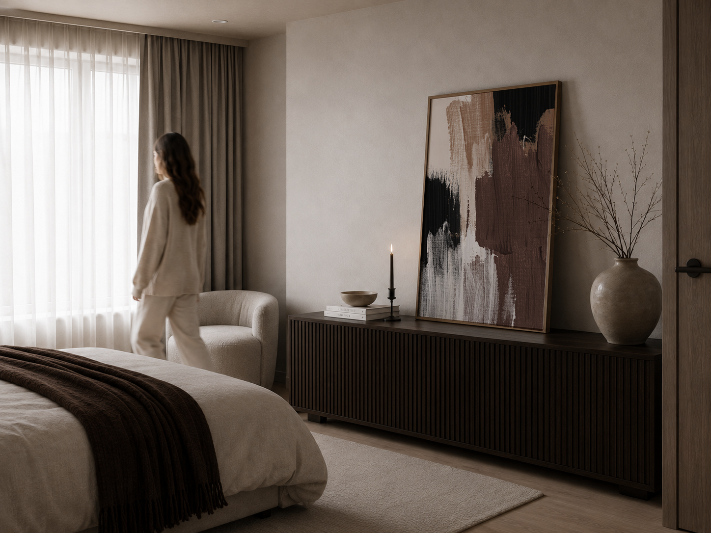 A woman in a cream pajama set stands by a window in a modern, softly lit bedroom. The room features a dark wood ribbed sideboard with abstract wall art, a lit candle, and a ceramic vase with dried branches.